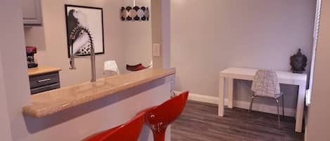Une cuisine moderne avec un comptoir en granit, des tabourets de bar rouges, une table à manger recouverte d’une nappe blanche et un luminaire fixé au mur.