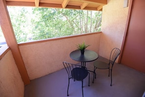 Uma pequena varanda com uma mesa redonda e duas cadeiras, um vaso de planta e vista para árvores através de uma janela.