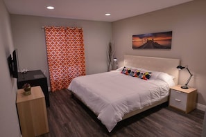 Um quarto com cama, mesa de cabeceira, luminária, televisão e um quadro na parede.