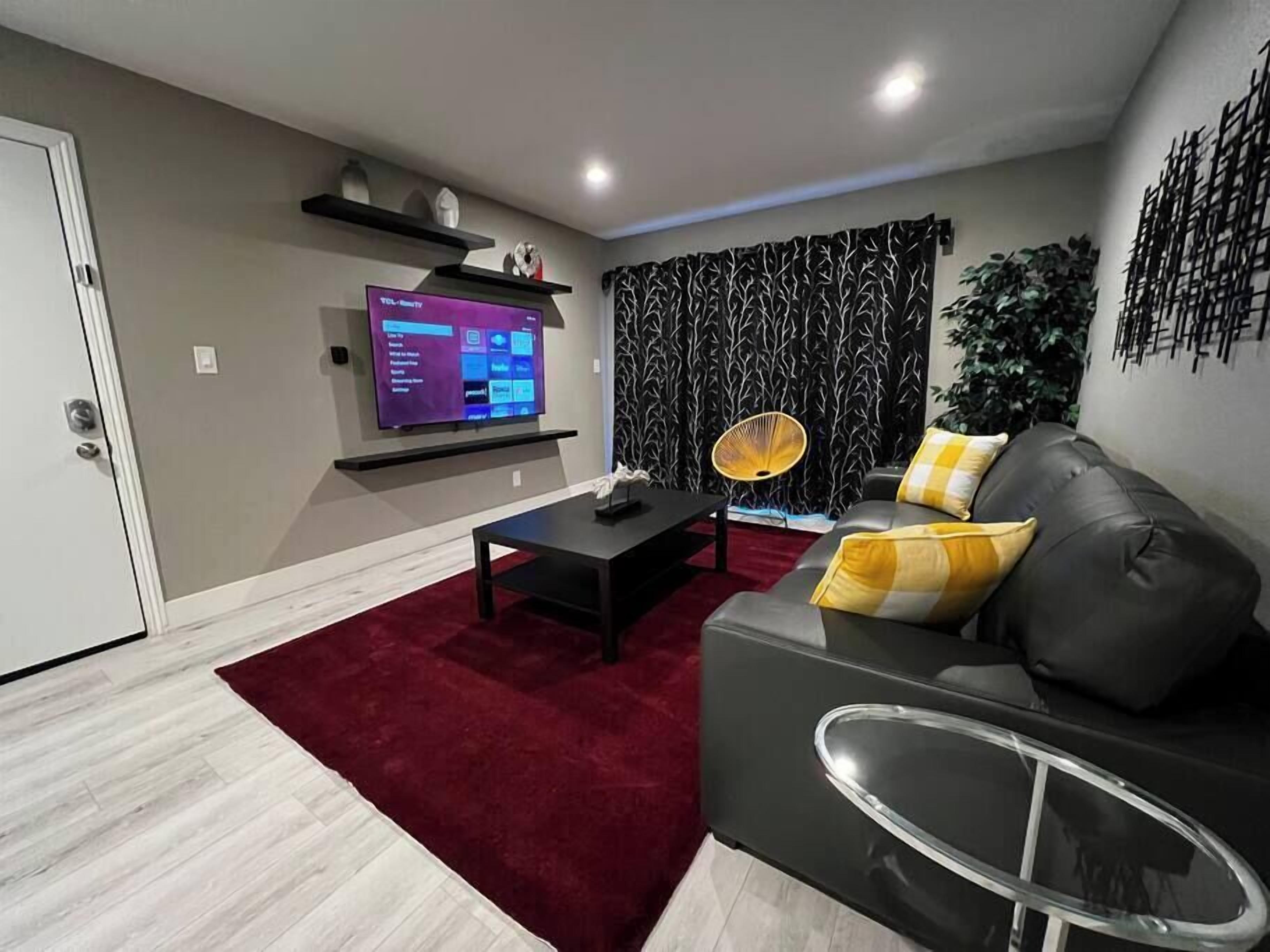 Un salon moderne avec un téléviseur à écran plat, un tapis rouge, un canapé noir avec des coussins jaunes et un élément décoratif mural.