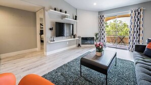 A modern living room with a flat-screen TV, a coffee table, and a sofa. There is a balcony with patterned curtains and a view of greenery. - Ruxley Suite Luxury 2b/2b condo (Las Vegas)
