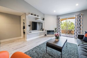 Ein modernes Wohnzimmer mit Fernseher, Couchtisch und Sofa. Ein Balkon mit gemusterten Vorhängen und Blick auf Grünflächen.