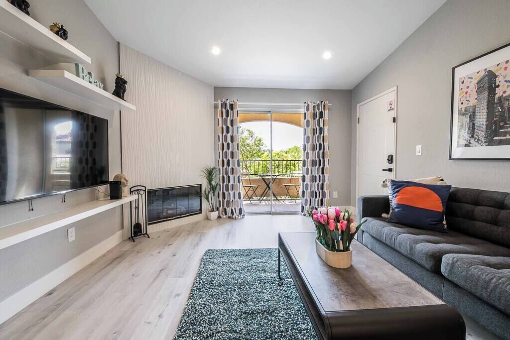 A modern living room with a flat-screen TV, a sofa, a coffee table with flowers, and a balcony with patterned curtains.