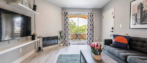 A modern living room with a flat-screen TV, a sofa, a coffee table with flowers, and a balcony with patterned curtains.