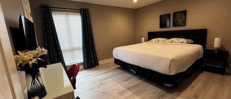 Un dormitorio con cama, escritorio, silla, ventana con cortinas y una obra de arte en la pared.