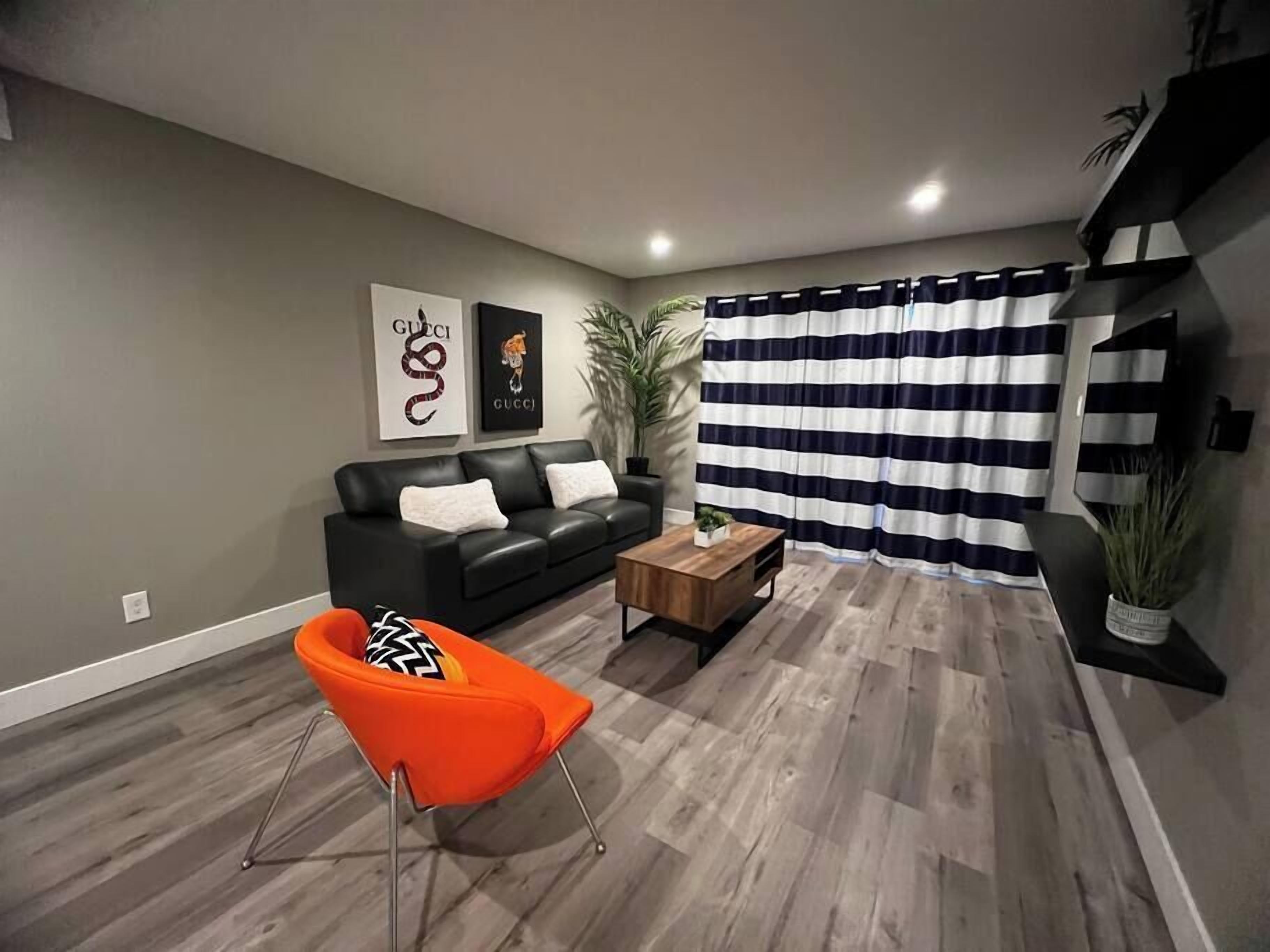 Een moderne woonkamer met een zwarte leren bank, een oranje fauteuil, een houten salontafel en een gestreept gordijn.