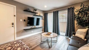 Een woonkamer met een flatscreen televisie, een ronde salontafel, een bank en een groot raam met patroon gordijnen.