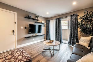 A living room with a flat-screen TV, a round coffee table, a sofa, and a large window with patterned curtains. - Glasgow Suite 1b/1b entire condo (Las Vegas)