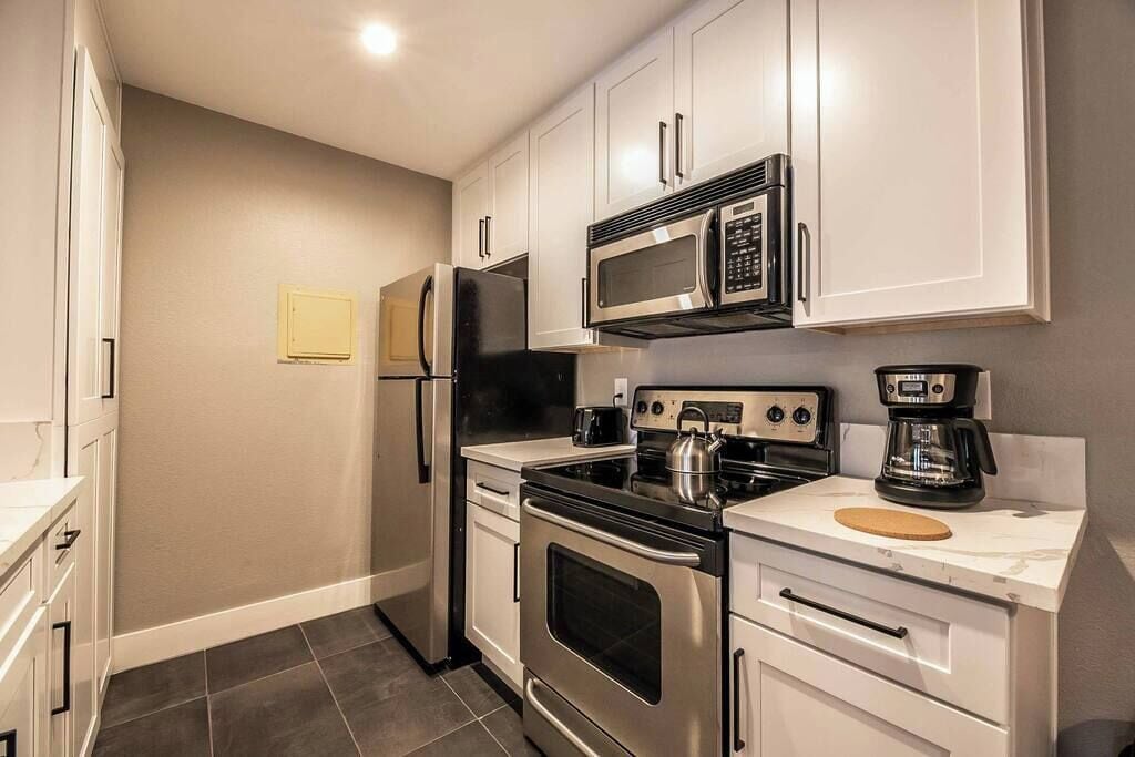 Una cocina compacta con gabinetes blancos, un refrigerador de acero inoxidable, un microondas, una estufa y una cafetera.