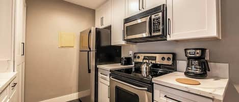 Una cocina compacta con gabinetes blancos, un refrigerador de acero inoxidable, un microondas, una estufa y una cafetera.