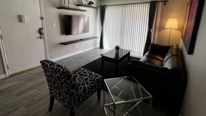 Ein gemütlicher Wohnraum mit Sofa, einem gemusterten Sessel, einem kleinen Tisch, einem an der Wand befestigten Fernseher und einem Fenster mit Vorhängen.