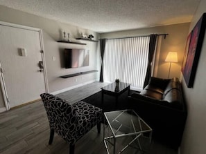 Ein gemütlicher Wohnraum mit Sofa, einem gemusterten Sessel, einem kleinen Tisch, einem an der Wand befestigten Fernseher und einem Fenster mit Vorhängen.