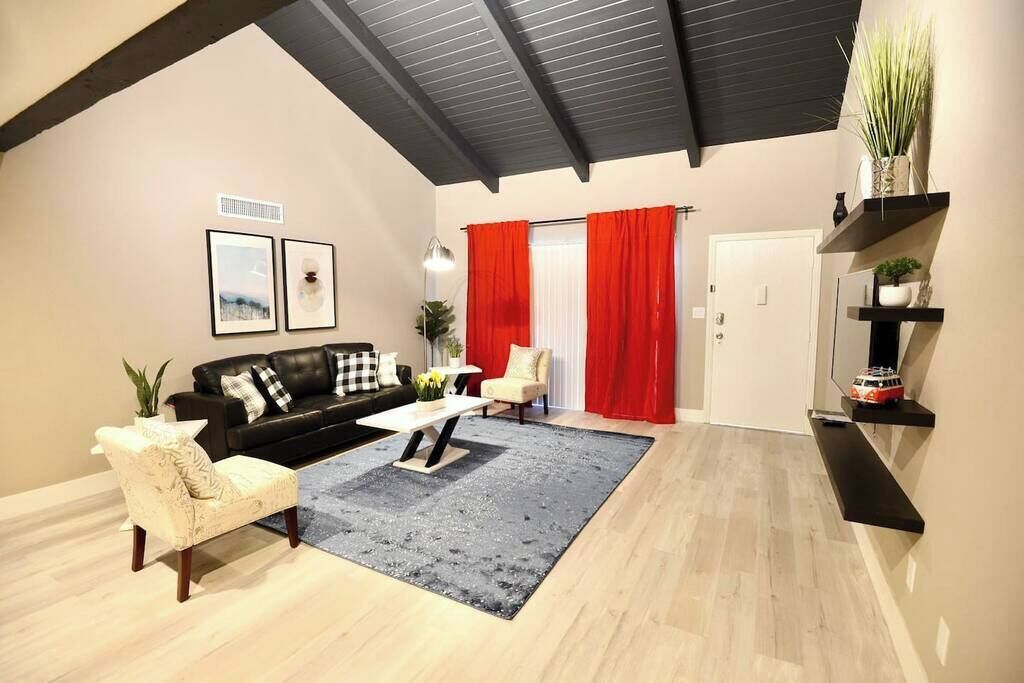 A living room with a black sofa, a white armchair, a coffee table, and a grey rug. There are red curtains, a door, and a shelf with plants.