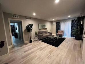 A modern living room with a sofa, coffee table, and a large plant. - Alpine Suite 2 Bed/1 Bath (Las Vegas)