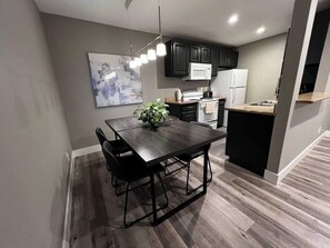 A modern kitchen with dark cabinetry, a wooden dining table, and a hanging light fixture. - Noma Suite Entire 2bed/2bath (Las Vegas)