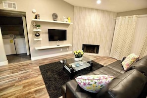 A modern living room with a leather sofa, a glass coffee table, a flat-screen TV mounted on a white wall, and a fireplace. - Chelsea Suite 2b/2b Strip View! (Las Vegas)