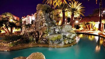 A nighttime resort pool area with a rock formation and palm trees.