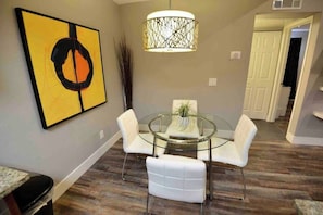 A dining area with a glass table and white chairs, a modern pendant light, and a piece of abstract art on the wall. - Chelsea Suite 2b/2b Strip View! (Las Vegas)