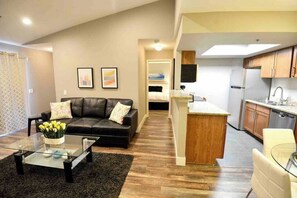 A modern living room with a glass coffee table, a black leather sofa, and a kitchen area with wooden cabinets and a built-in island. - Chelsea Suite 2b/2b Strip View! (Las Vegas)