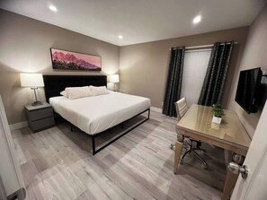 Ein Hotelzimmer mit Bett, Nachttisch, Lampe, einem Bild an der Wand, einem Fenster mit Vorhängen und einem Fernseher.