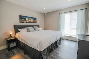 A hotel room with a bed, a nightstand, a lamp, a window with curtains, and a picture on the wall. - Sundance Suite 2B/2B Central (Las Vegas)