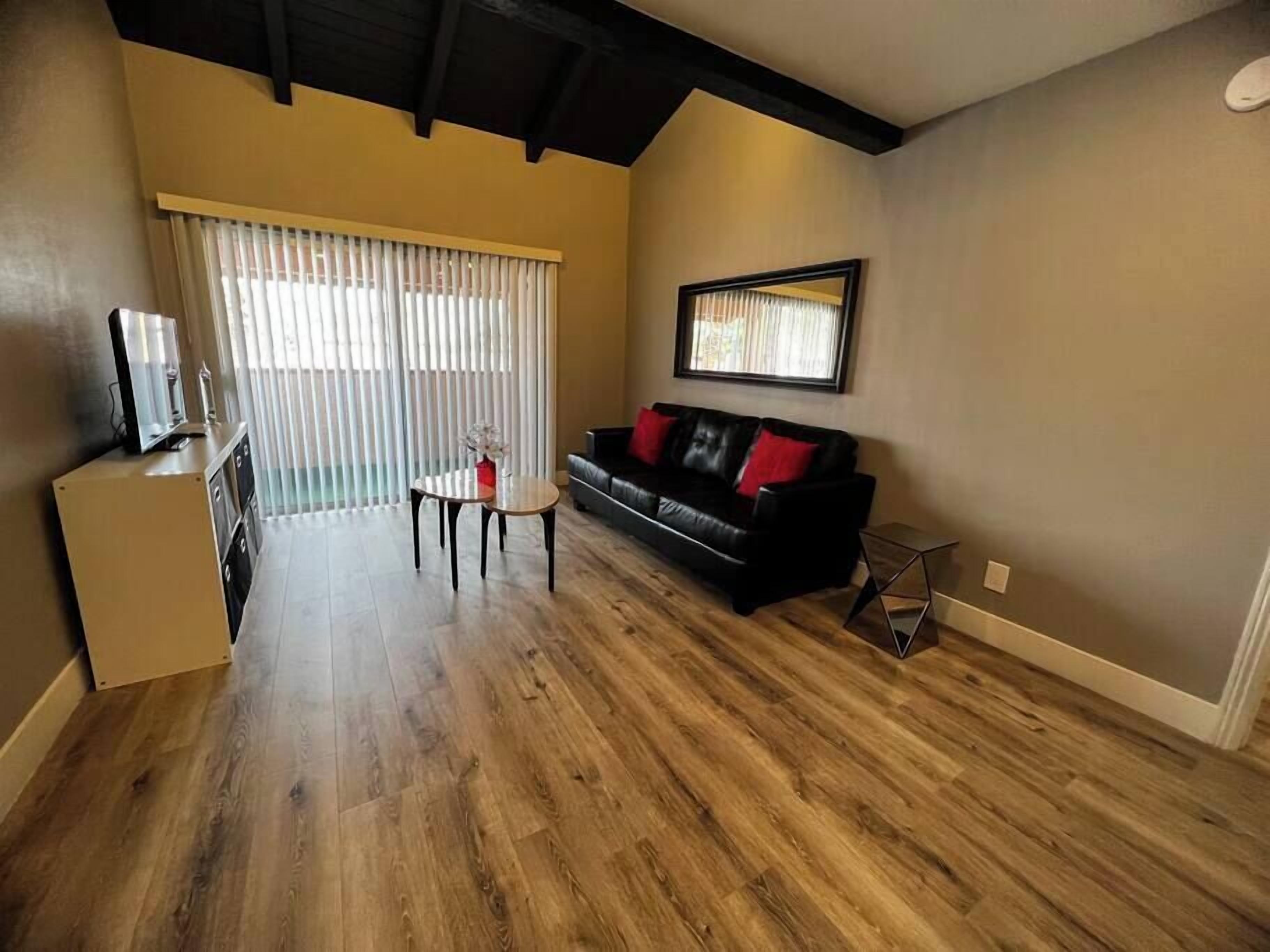 A living room with a wooden floor, a black sofa with red cushions, a small table, and a window with blinds.