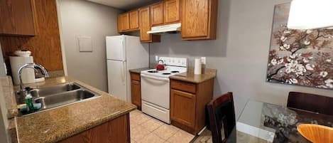 Una cocina funcional con armarios de madera, un refrigerador blanco, una estufa y una zona de comedor con una mesa de vidrio.