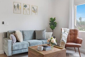 Een woonkamer met een lichtblauwe bank, een houten salontafel en een bruine leren fauteuil. Op de muur hangen ingelijste botanische prints en in de hoek staat een grote groene plant.