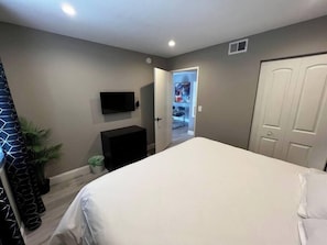 Ein Schlafzimmer mit einem Bett, einem an der Wand montierten Fernseher, einer Tür und einer kleinen Pflanze.