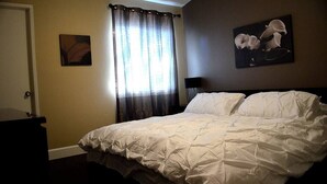 Un dormitorio con una cama, una puerta, un ventanal con cortinas y dos obras de arte colgadas en la pared.