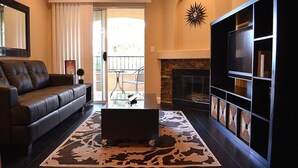 Una sala de estar con un sofá de cuero oscuro, un televisor de pantalla plana, una chimenea y un ventanal con cortinas.