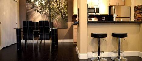 A modern kitchen with stainless steel appliances, a dining area with black chairs, and a bar stool area.