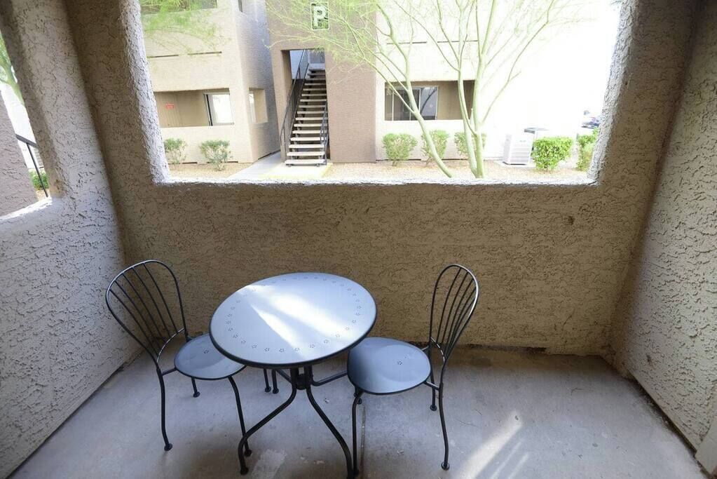 En lille patio med et rundt bord og to stole, et vindue med udsigt til en bygning og et træ, samt en beton gulv.