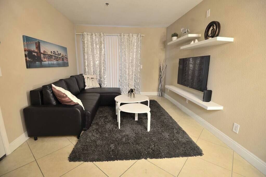 En stue med en sort sofa, et lille hvidt bord, en mørk måtte, et fjernsyn på en hylde og et billede på væggen.