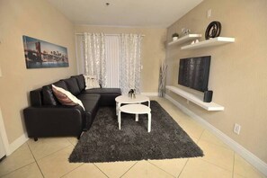 A living room with a black sofa, a small white table, a dark rug, a TV on a shelf, and a painting on the wall. - Ava Suite 1 B/1B close to strip (Las Vegas)
