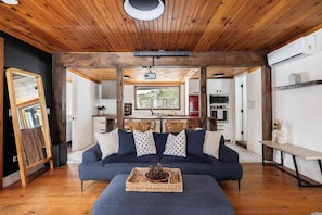 Ein gemütliches Wohnzimmer mit einem dunkelblauen Sofa, Holzbalkendecke und einer Küche im Hintergrund.