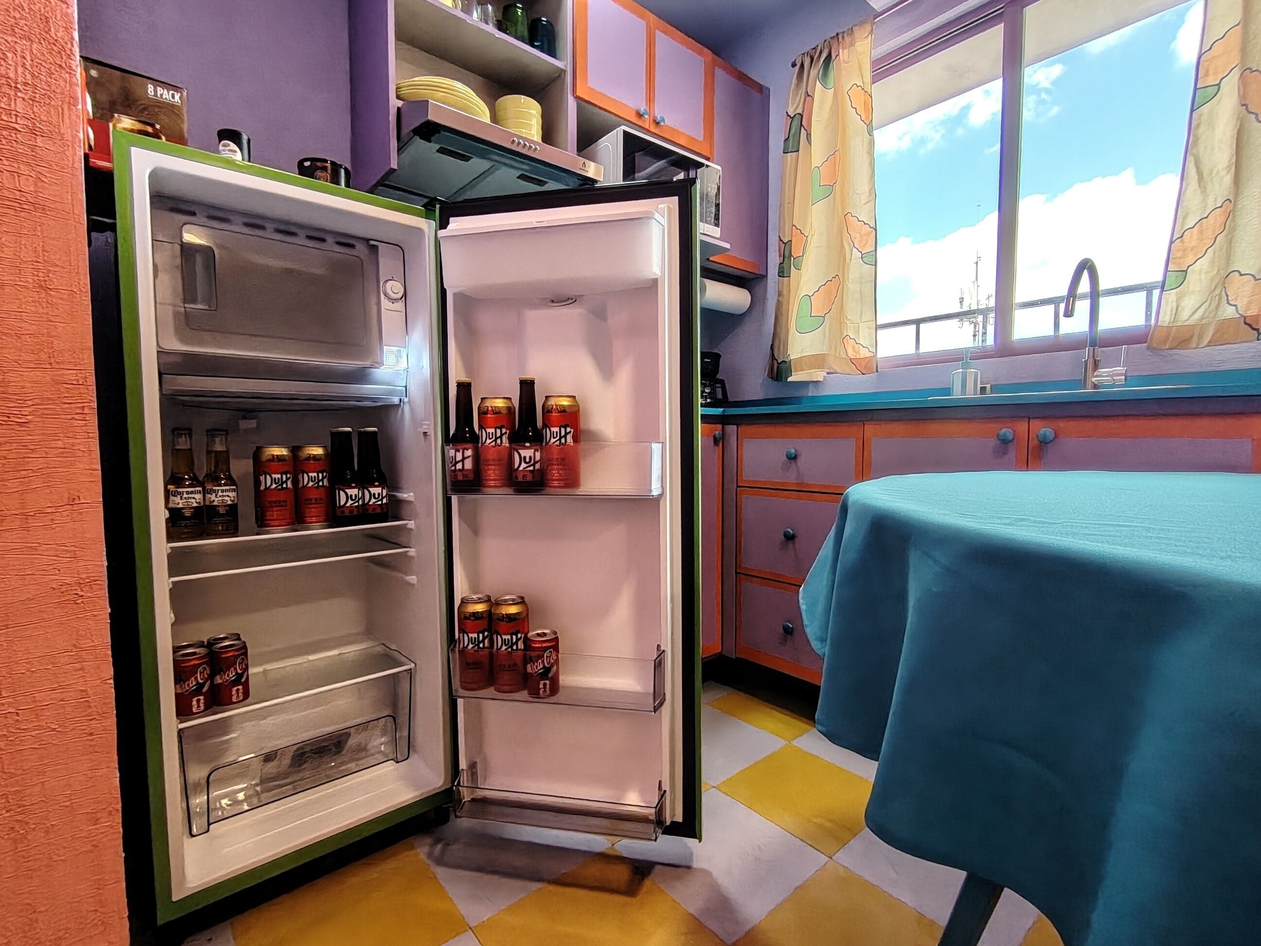 Uma cozinha com geladeira repleta de bebidas, uma janela com vista e uma mesa com toalha azul.