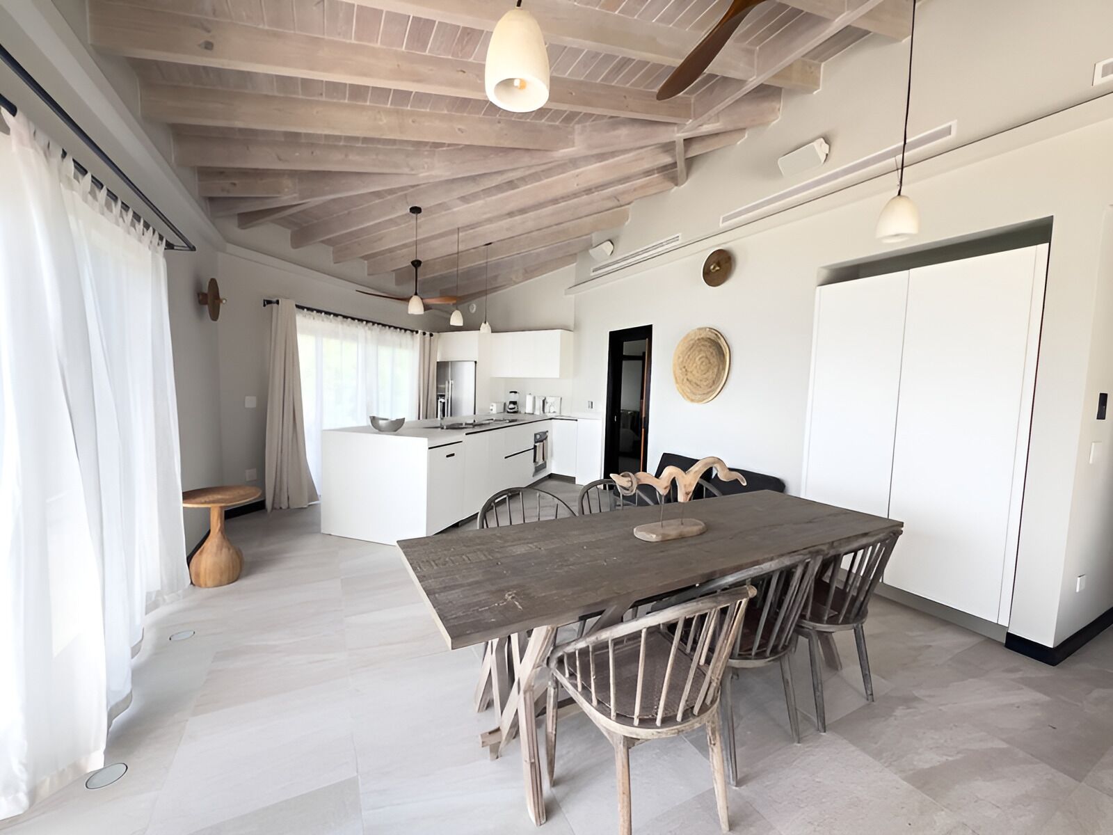 Un vaste espace de vie comprenant une grande table à manger et des chaises en bois, une cuisine à l’arrière-plan et un plafond avec des poutres apparentes.