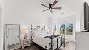 A bedroom with a bed, nightstands, a ceiling fan, a window with a view, and a door. - Magnolia Pointe 4893-304 (Myrtle Beach)