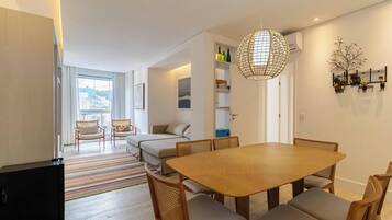 A modern living room with a wooden dining table, cushioned chairs, a sofa, and a large window.
