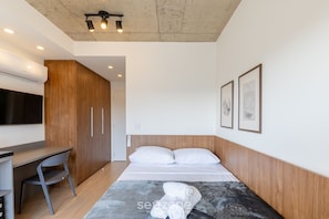 Een moderne hotelkamer met een houten garderobe, een bureau, een bed met witte lakens en twee ingelijste kunstwerken aan de muur.