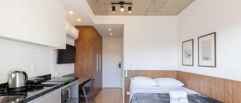 Una habitación de hotel compacta con kitchenette, una cama con manta gris y dos obras de arte enmarcadas.