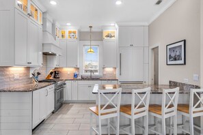 A modern kitchen with white cabinetry, a central island with bar stools, and a window with blinds. - Sea Glass, Beach Front, Renovated, Community Pools, Beach Gear Credit! (St. George Island)