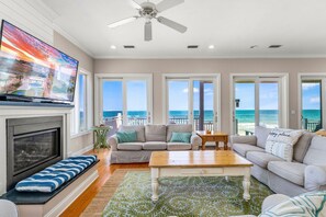 A spacious living room with a fireplace, comfortable seating, a television, and large windows offering ocean views. - Sea Glass, Beach Front, Renovated, Community Pools, Beach Gear Credit! (St. George Island)