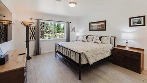 Ein Schlafzimmer mit Bett, Nachttischen, einem Schminktisch, einem Fenster mit Vorhängen und Blick auf Bäume und einen See.