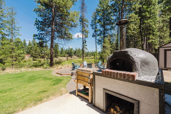 A wood-fired pizza oven with a chimney, surrounded by a grassy area, trees, and outdoor seating.