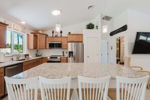 Cocina moderna con encimeras de granito, gabinetes de madera y electrodomésticos de acero inoxidable, además de una isla grande con sillas blancas.
