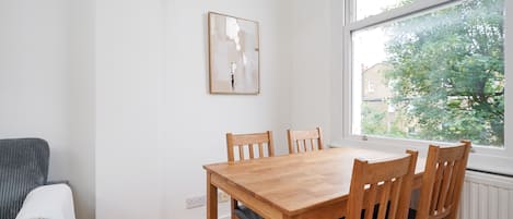 Un coin repas avec une table et des chaises en bois, une fenêtre donnant sur des arbres et un mur blanc orné d’un tableau encadré.