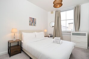 Ein Hotelzimmer mit einem Bett, zwei Nachttischlampen, einem Nachttisch, einem Heizkörper, einem Fenster mit Vorhängen und einem an der Wand befestigten Spiegel.