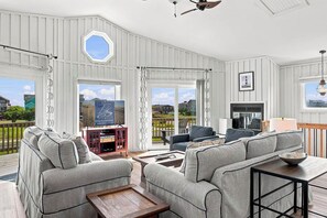 A spacious living room with a large sofa set, a coffee table, and a fireplace. There are large windows offering a view of the outdoors. - Carolina Sol | 560 ft to the beach | Private Pool and Community Pool (Nags Head)